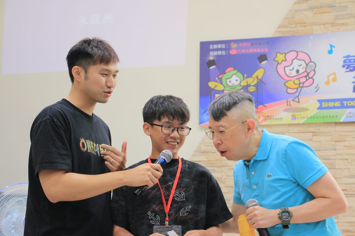 成果發表會上，學員手持麥克風上台分享，導師在一旁陪伴。